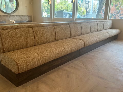 Long upholstered booth seating with brown cushions and dark base, positioned near windows with wooden flooring
