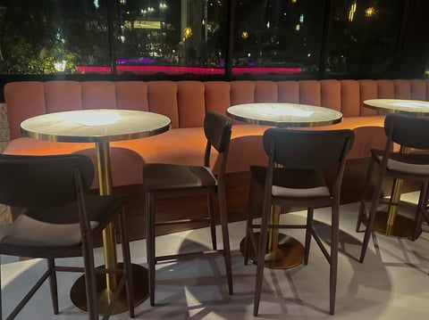 Modern outdoor lounge seating area with high-top tables, orange upholstered booths, dark chairs, and city skyline at night