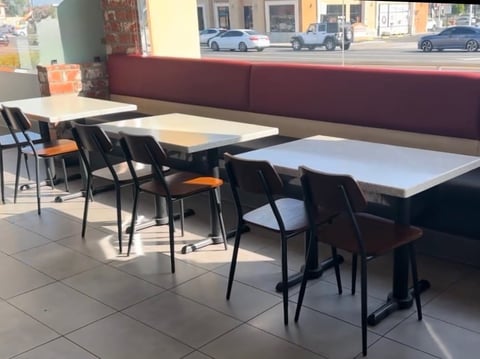 Modern café interior with dining tables and black chairs beside a burgundy banquette, street view visible through large windows