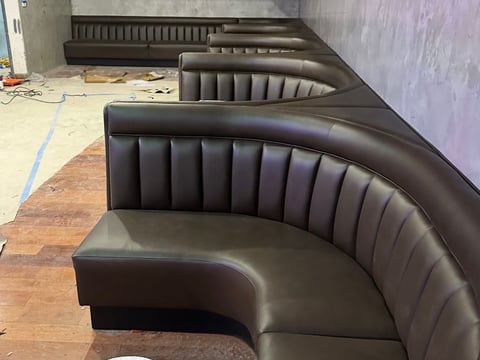 Black leather curved booth seating with vertical channel quilting under construction in an empty room with concrete walls and wooden flooring.