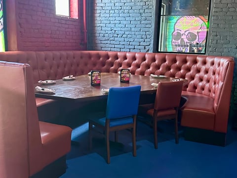 Restaurant booth with red tufted seating, blue and red chairs around a table with condiments and neon skull artwork on brick walls.