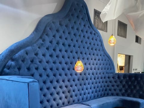 Large blue quilted oversized armchair with tufted details displayed in a bright architectural courtyard with pendant lights.