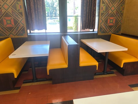 Modern restaurant booths with yellow seats and white tables, separated by dividers, with geometric patterned wallpaper and large window in background